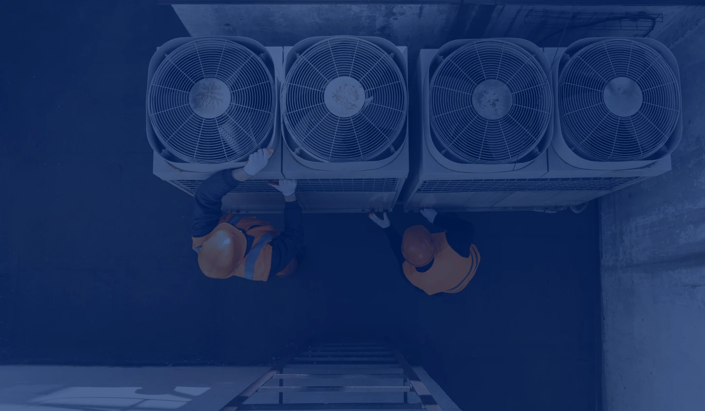 Two workers in orange safety vests and helmets inspecting four large rooftop air conditioning units.