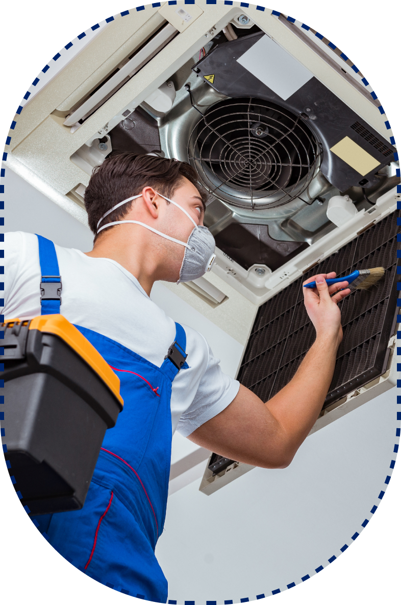 A masked technician in blue overalls repairs a ceiling AC, holding a toolbox and using a screwdriver.
