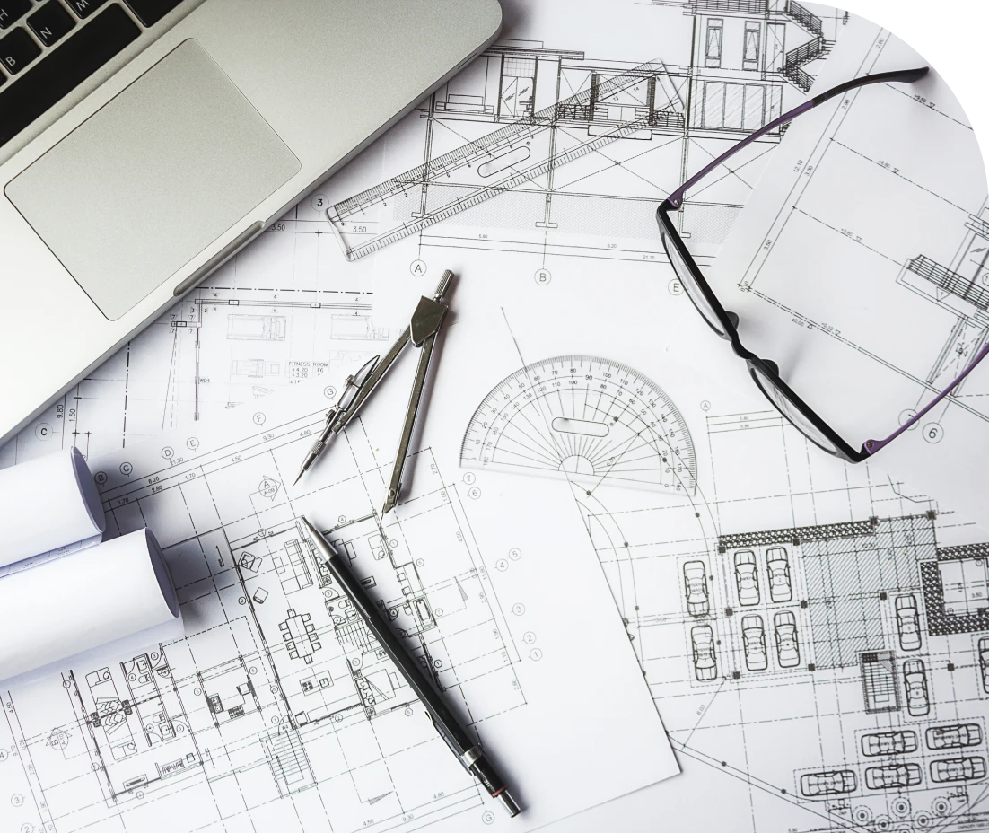 Architectural plans on a table with a laptop, rolled blueprints, a protractor, a compass, glasses.